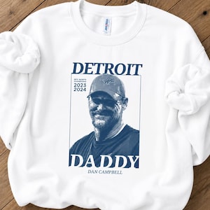 May include: A white crewneck sweatshirt featuring a dark blue graphic print. The design includes the text DETROIT, DADDY, and DAN CAMPBELL, along with the words NFC NORTH CHAMPIONS 2023 2024 and a portrait of a man wearing a baseball cap.