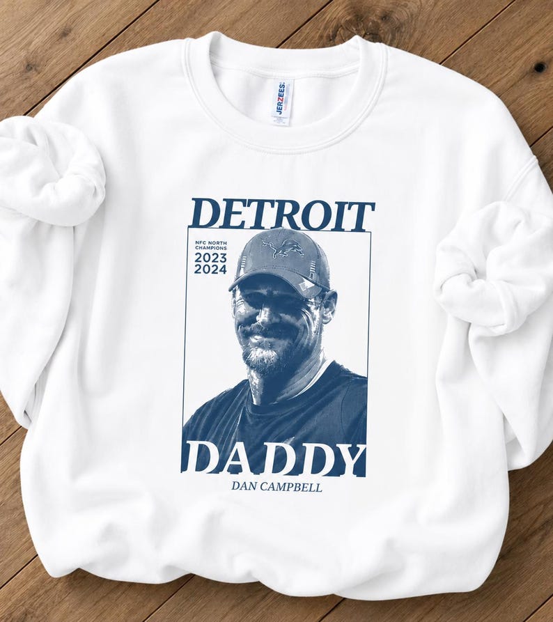 May include: A white crewneck sweatshirt featuring a dark blue graphic print. The design includes the text DETROIT, DADDY, and DAN CAMPBELL, along with the words NFC NORTH CHAMPIONS 2023 2024 and a portrait of a man wearing a baseball cap.