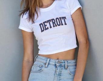 May include: A white short-sleeved crop top t-shirt featuring the word DETROIT printed in bold navy blue letters with an orange outline across the chest, paired with light blue denim jeans that have a visible metal button and zipper fly closure.