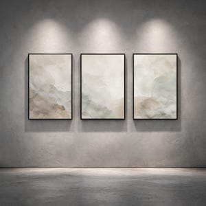 May include: Three framed abstract art prints. Each print features a watercolour-style design in neutral tones of beige, grey, and brown. The frames are black, and the artwork is displayed on a grey wall with spotlights.