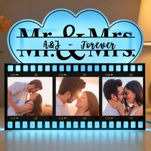 Peut inclure: Une enseigne lumineuse en forme de nuage avec un éclairage bleu portant le texte Mr. & Mrs. A&J - Forever. Sous l'enseigne se trouve un cadre en forme de pellicule photo contenant trois photos d'un couple, avec des bords noirs et des perforations.