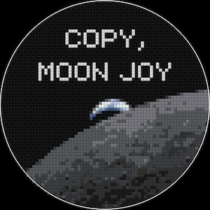 May include: A cross-stitch design in a black circular frame. The text "COPY, MOON JOY" is stitched in white. Below the text is a pixelated depiction of the moon's surface with a small, blue and white spacecraft.