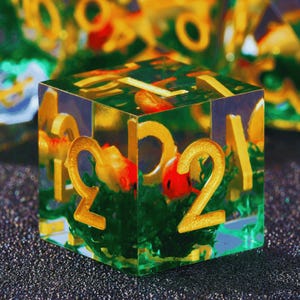 Puede incluir: Un dado de resina transparente con números dorados y una escena de jardín en miniatura en el interior. El dado presenta una base verde con detalles en rojo y amarillo, creando un accesorio de juego único.