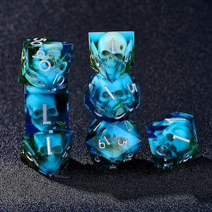 Puede incluir: Un conjunto de seis dados con diseño de calavera. Los dados son azules y verdes, con números plateados. Las calaveras son visibles dentro de los dados translúcidos. Los dados están apilados y dispersos.