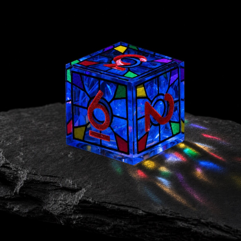 May include: A translucent blue cube-shaped die featuring a stained-glass pattern with red, yellow, and green accents. The faces display large red numbers, including a six and a two, with light refracting through the material to create colorful patterns.