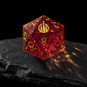 May include: A twenty-sided die featuring a translucent deep red resin body with intricate black geometric line patterns and gold-colored numbers. The top face displays a stylized gold emblem of a three-pronged crest instead of a number.
