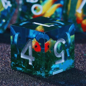 May include: A clear resin die with a miniature underwater scene. The die features two orange and yellow fish, green plant life, and the numbers 4 and 5 in silver. The background is a mix of blue and green.
