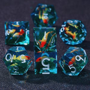Puede incluir: Conjunto de seis dados azules y verdes con un diseño de pez dorado. Los dados están numerados del 1 al 6.