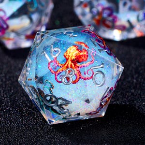 May include: A clear, multi-sided die with a vibrant red and orange octopus design suspended within. The die features silver numbering and is filled with blue glitter, creating a fantasy-themed aesthetic.
