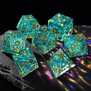 May include: A set of seven polyhedral dice featuring a translucent teal and blue stained glass pattern with gold-colored numbers and metallic lead-like lines. The dice have various geometric shapes including cubes, pyramids, and multi-faceted forms.