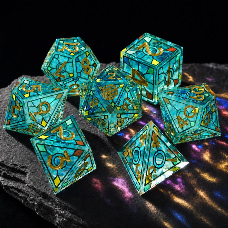 May include: A set of seven polyhedral dice featuring a translucent teal and blue stained glass pattern with gold-colored numbers and metallic lead-like lines. The dice have various geometric shapes including cubes, pyramids, and multi-faceted forms.