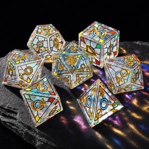 May include: A set of seven polyhedral dice featuring a clear, iridescent resin base with a stained-glass style pattern of black lines and colorful geometric shapes. Each die has gold-colored numbers etched into the surface, reflecting light.