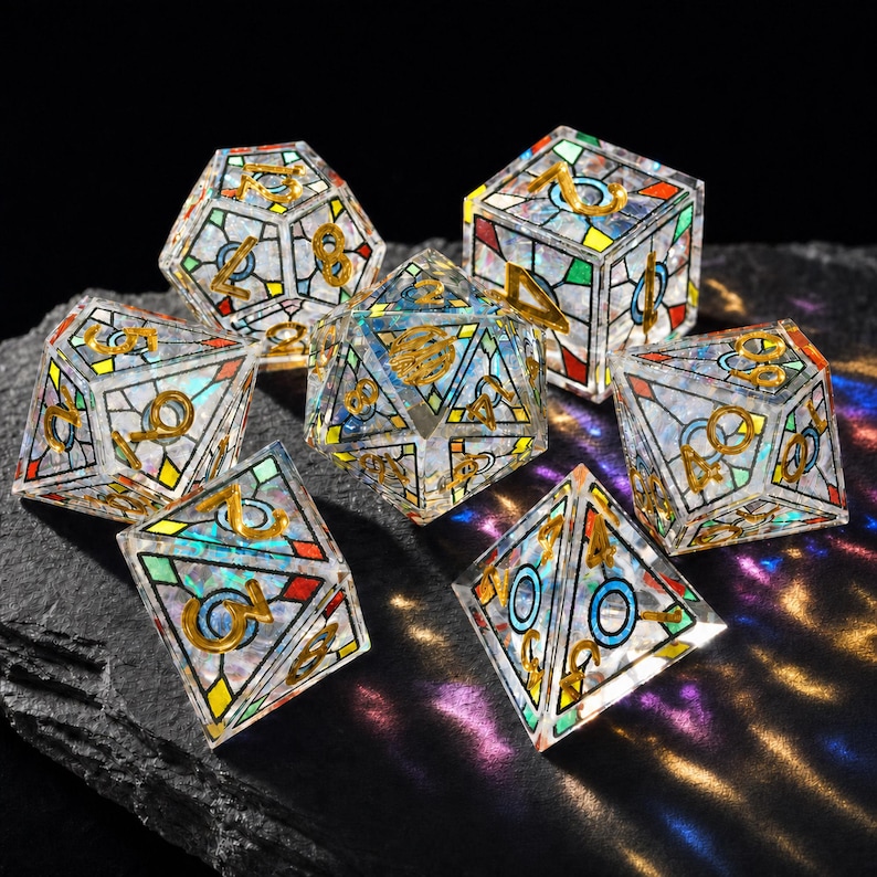 May include: A set of seven polyhedral dice featuring a clear, iridescent resin base with a stained-glass style pattern of black lines and colorful geometric shapes. Each die has gold-colored numbers etched into the surface, reflecting light.