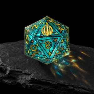 May include: A twenty-sided die featuring a translucent teal and blue stained glass pattern with gold-colored geometric lead lines and gold-colored numbers. The die displays a prominent gold symbol at the top and is resting on a dark, textured surface.