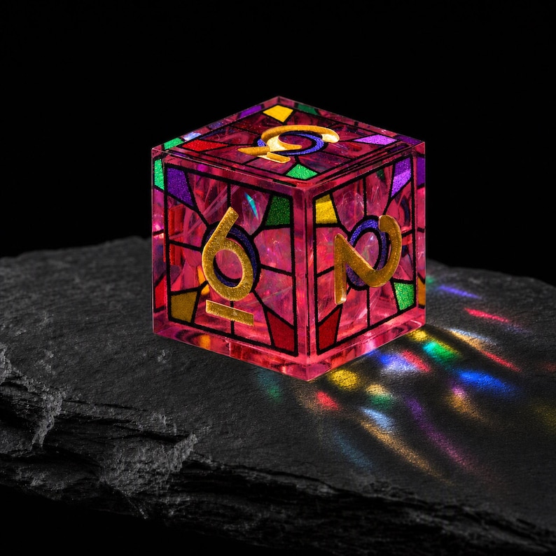 May include: A six-sided die featuring a stained glass aesthetic with a translucent pink resin body. The faces are decorated with black grid lines and multi-colored geometric shapes in green, yellow, and purple. Gold numerals are embossed on the faces.
