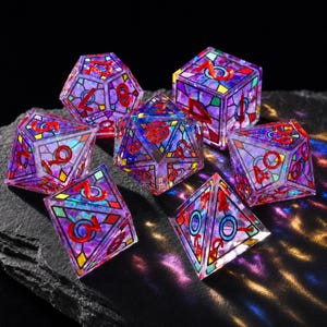 May include: A set of seven polyhedral dice featuring a stained-glass aesthetic with translucent purple, blue, and green panels outlined in black. Each die has bright red numbers embossed on the faces, creating a vibrant, multi-faceted appearance.
