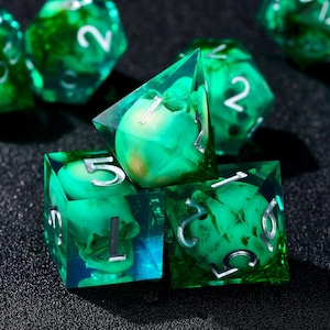 May include: A set of green and clear resin dice with silver numbering. The dice have a unique design with embedded green and yellow elements, creating a layered effect. The dice are displayed on a dark surface.