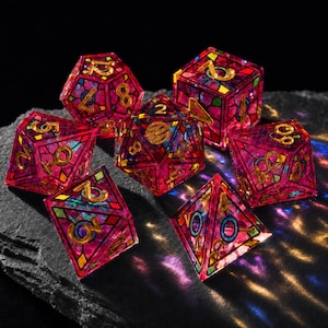 May include: A set of seven polyhedral dice featuring a translucent magenta resin base with a stained-glass style grid pattern in black, gold, and teal. Each die face displays gold-colored numbers, with some faces showing circular accents.