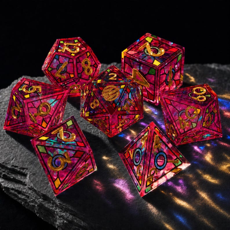 May include: A set of seven polyhedral dice featuring a translucent magenta resin base with a stained-glass style grid pattern in black, gold, and teal. Each die face displays gold-colored numbers, with some faces showing circular accents.