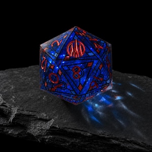 May include: A twenty-sided die featuring a deep blue translucent body with a stained glass-like grid pattern. The numbers are engraved in a metallic red finish, with a prominent red symbol replacing the number twenty on the top face.