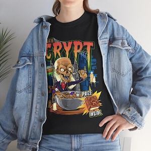 May include: Black t-shirt with a graphic of a zombie eating cereal from a bowl. The word "CRYPT" is in large orange letters above the zombie. The text "PRIZE INSIDE" is below a book. The shirt is worn with a blue denim jacket.
