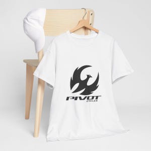 May include: White t-shirt with a black graphic of a stylized bird and the text "PIVOT CYCLES" below it. A white baseball cap hangs on a wooden chair. The t-shirt is hanging on a wooden hanger.