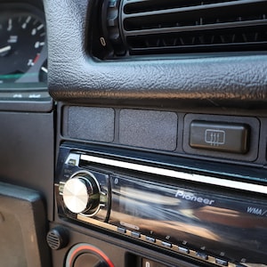 Puede incluir: Primer plano del salpicadero de un coche con un sistema estéreo Pioneer. El salpicadero negro tiene una superficie texturizada, rejillas de ventilación, un botón de desempañado y dos paneles rectangulares en blanco. El estéreo tiene un mando plateado y una pantalla digital.