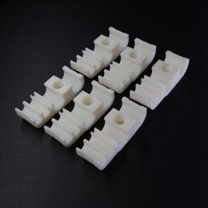 Könnte beinhalten: Sechs cremefarbene Kunststoffkomponenten mit rechteckiger Basis, einem zentralen Loch und Rillen. Sie sind auf einer dunklen Oberfläche angeordnet.