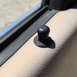 May include: A black car door lock pin with a round top and a textured base. The pin is set against a beige, textured car door panel. A blue car door frame is visible in the background.