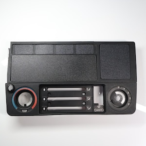 Puede incluir: Componente de salpicadero de coche de plástico negro con varios controles. Incluye un dial de temperatura con marcas azules y rojas, controles de ventilación y un dial de velocidad del ventilador. Superficie texturizada y varios compartimentos rectangulares.