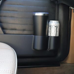 May include: Black car door cup holder with a black tumbler and a silver and white Zero Sugar Monster Energy drink can. The cup holder is integrated into the door panel.