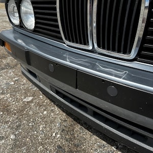 May include: Close-up of a classic car's front end. The image showcases the black grill, headlights, and bumper. The bumper has two black sensors and a silver trim. The car is parked on a textured, gray surface.