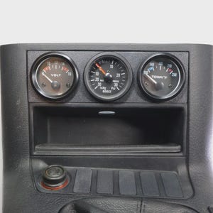 以下が含まれることがあります： 3つの丸いゲージが付いた黒いダッシュボードパネル。ゲージは、電圧、真空/ブースト圧、華氏温度を表示します。パネルには、小さな収納スペースとシガレットライターも付いています。