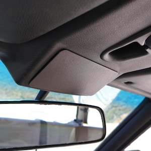 以下が含まれることがあります： 車の内装のクローズアップ。バックミラーとオーバーヘッドコンソールに焦点を当てています。コンソールには長方形のグレーのサンバイザーがあり、テクスチャ加工された濃いグレーの表面に取り付けられています。バックミラーは黒です。