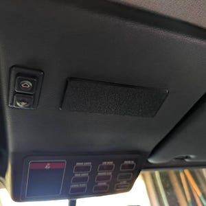Puede incluir: Panel interior negro del techo del coche con varios botones de control. Incluye botones para cerraduras de puertas, luces y otras funciones del vehículo. También se ve un parasol.