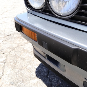 May include: Close-up of a classic car's front end. The image shows a gray bumper with an amber side marker light and a black trim. Two round headlights are visible above the bumper. The car is parked on a cracked concrete surface.