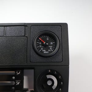 Könnte beinhalten: Schwarzes Kfz-Armaturenbrett-Bauteil mit einem runden Messgerät. Das Messgerät hat ein schwarzes Zifferblatt, weiße Markierungen und einen roten Zeiger, der Vakuum- und Ladedruck anzeigt. Die Einheit zeigt inHg und Psi an. Weitere Bedienelemente und Lüftungsschlitze sind sichtbar.