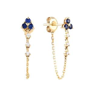 May include: A pair of gold earrings with a cluster of three blue gemstones at the top. Below the cluster, a delicate chain suspends three smaller white gemstones. The earrings have a post and chain design.
