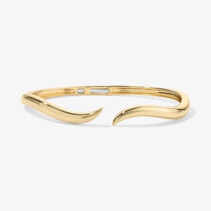 May include: A gold-colored bangle bracelet with a unique, open design. The bracelet has a smooth, curved shape with a gap at the front. The ends of the bracelet curve inward, creating a stylish and modern look. The bracelet is made of a shiny, reflective metal.