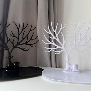 May include: Two decorative jewelry stands. One is black, the other white. Each features a deer base with a tree-like structure for hanging necklaces and bracelets. The stands sit on oval trays, offering a stylish way to organize accessories.