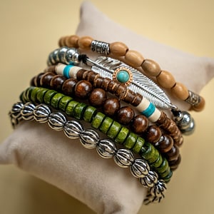 May include: A stack of beaded bracelets in various styles and colors. The bracelets include wooden beads, silver-colored metal beads, and green beads. One bracelet features a silver feather charm with a turquoise stone.
