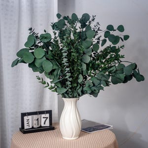May include: A white ceramic vase with a wavy design holds a bouquet of green eucalyptus branches. The vase is sitting on a light brown table with a black calendar that reads "JAN 17".