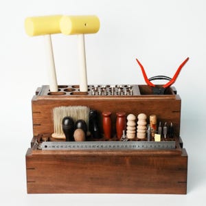 May include: A dark brown wooden tool organizer stand holding various leatherworking tools, including two yellow mallets, a brush, wooden burnishers, metal punches, and red-handled pliers, with a 30 cm metal ruler placed across the front.