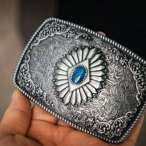 May include: Silver-coloured rectangular belt buckle with detailed floral and scroll patterns. The buckle has a central design with a blue gemstone. The edges have a detailed rope-like border.