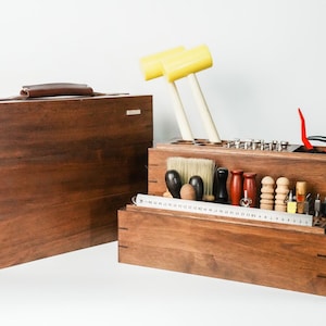 May include: A dark brown wooden tool storage case with a handle and a matching open organizer box containing various leatherworking tools, including two yellow mallets, a metal ruler, brushes, and assorted wooden and metal hand tools.