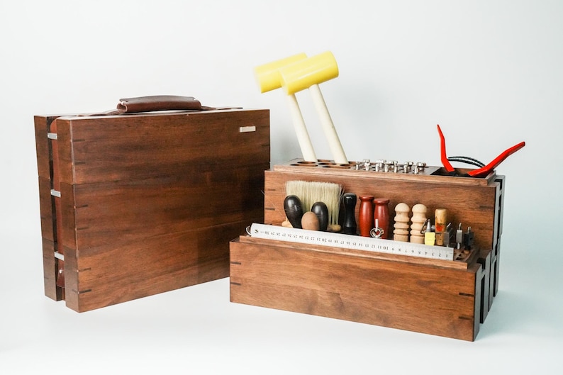 May include: A dark brown wooden tool storage case with a handle and a matching open organizer box containing various leatherworking tools, including two yellow mallets, a metal ruler, brushes, and assorted wooden and metal hand tools.