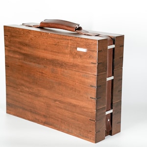 May include: A rectangular wooden carrying case with a dark brown finish, featuring visible joinery on the edges. The case includes a brown leather handle on top and a matching leather strap secured by silver-toned metal hardware on the side.