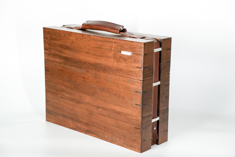 May include: A rectangular wooden carrying case with a dark brown finish, featuring visible joinery on the edges. The case includes a brown leather handle on top and a matching leather strap secured by silver-toned metal hardware on the side.