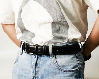 May include: A black leather belt with a silver buckle is worn with light blue denim jeans. The belt is secured around the waist. The person is wearing a white t-shirt with a large grey design.