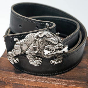 May include: A black leather belt with a silver bulldog buckle. The bulldog is holding a bone in its mouth.
